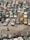 An aerial view of an outdoor area filled with numerous old and rusted cars parked closely together. The cars display a range of colors and visible signs of decay and neglect, such as peeling paint and rust. A person wearing dark clothing and a hat is walking on the rocky ground near the bottom of the image.