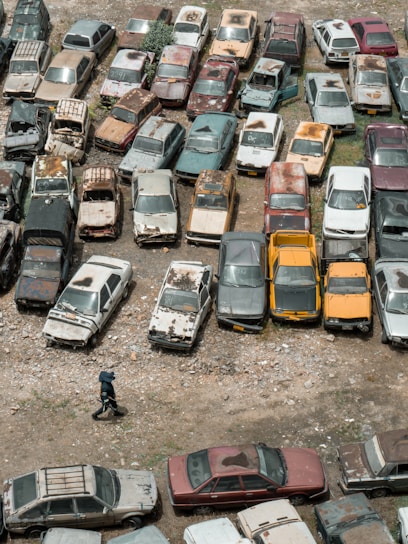 An aerial view of an outdoor area filled with numerous old and rusted cars parked closely together. The cars display a range of colors and visible signs of decay and neglect, such as peeling paint and rust. A person wearing dark clothing and a hat is walking on the rocky ground near the bottom of the image.