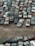 An overhead view of numerous old and rusted cars tightly packed together in a junkyard. The vehicles exhibit significant wear and tear, with visible rust, dents, and peeling paint. The land between the cars is sparse with rocks and patches of dry grass.