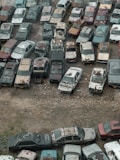 An overhead view of numerous old and rusted cars tightly packed together in a junkyard. The vehicles exhibit significant wear and tear, with visible rust, dents, and peeling paint. The land between the cars is sparse with rocks and patches of dry grass.