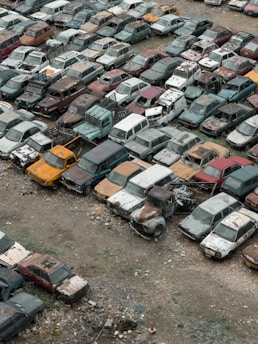 A large collection of old, rusting cars is tightly packed in an outdoor junkyard. The vehicles are in various states of disrepair, with peeling paint and visible rust. The ground around the cars is covered with dirt and scattered debris.