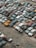 A large collection of old, rusting cars is tightly packed in an outdoor junkyard. The vehicles are in various states of disrepair, with peeling paint and visible rust. The ground around the cars is covered with dirt and scattered debris.