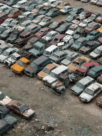 A large collection of old, rusting cars is tightly packed in an outdoor junkyard. The vehicles are in various states of disrepair, with peeling paint and visible rust. The ground around the cars is covered with dirt and scattered debris.