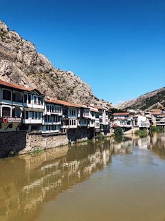 A row of traditional, Ottoman-style houses sit along a riverbank, reflecting in the calm water. The buildings are characterized by white facades and dark wooden frames. A rocky mountain dominates the background under a clear blue sky.