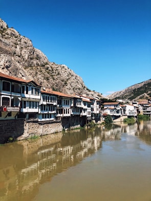 A row of traditional, Ottoman-style houses sit along a riverbank, reflecting in the calm water. The buildings are characterized by white facades and dark wooden frames. A rocky mountain dominates the background under a clear blue sky.