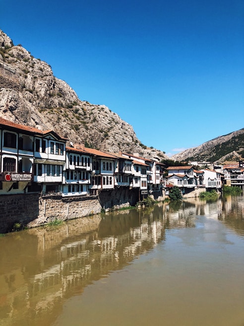 A row of traditional, Ottoman-style houses sit along a riverbank, reflecting in the calm water. The buildings are characterized by white facades and dark wooden frames. A rocky mountain dominates the background under a clear blue sky.