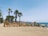 Large sand-colored sculptural letters on a sandy beach spell out 'Malagueta' with palm trees lining the background. People relax on the beach, with a vibrant yellow umbrella providing shade. The sea and clear, blue sky stretch out into the horizon.