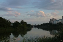 A serene river scene with greenery lining both banks, reflecting the soft hues of the evening sky filled with scattered clouds. On the right side, there are residential buildings in the distance, adding an urban touch to the natural landscape.