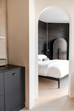 A modern bedroom with a minimalist design features a white bed with a wooden frame and a dark wooden headboard. The room is accessed through a rounded archway. A small black unit is present in the foreground on the left side. The walls are a mix of white and wood paneling. The lighting is soft, providing a calm ambiance.