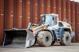 A heavy construction vehicle with large, rugged tires and a front loader bucket. The vehicle is parked on a dirt surface in front of a large, rust-colored metal wall. It has a predominantly yellow and gray color scheme and appears to be built for industrial use.