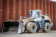 A heavy construction vehicle with large, rugged tires and a front loader bucket. The vehicle is parked on a dirt surface in front of a large, rust-colored metal wall. It has a predominantly yellow and gray color scheme and appears to be built for industrial use.