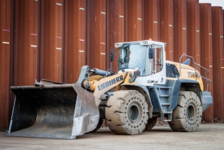 A heavy construction vehicle with large, rugged tires and a front loader bucket. The vehicle is parked on a dirt surface in front of a large, rust-colored metal wall. It has a predominantly yellow and gray color scheme and appears to be built for industrial use.