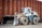A heavy construction vehicle with large, rugged tires and a front loader bucket. The vehicle is parked on a dirt surface in front of a large, rust-colored metal wall. It has a predominantly yellow and gray color scheme and appears to be built for industrial use.