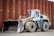 A heavy construction vehicle with large, rugged tires and a front loader bucket. The vehicle is parked on a dirt surface in front of a large, rust-colored metal wall. It has a predominantly yellow and gray color scheme and appears to be built for industrial use.