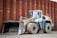 A heavy construction vehicle with large, rugged tires and a front loader bucket. The vehicle is parked on a dirt surface in front of a large, rust-colored metal wall. It has a predominantly yellow and gray color scheme and appears to be built for industrial use.