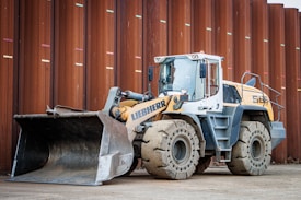 A heavy construction vehicle with large, rugged tires and a front loader bucket. The vehicle is parked on a dirt surface in front of a large, rust-colored metal wall. It has a predominantly yellow and gray color scheme and appears to be built for industrial use.