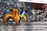 A large yellow mechanical claw is positioned in front of a massive pile of assorted metal scrap and debris. The ground is wet, suggesting recent rain, and the background shows a corrugated metal fence. Various pieces of metal, including bicycle parts and wires, are scattered and piled haphazardly.