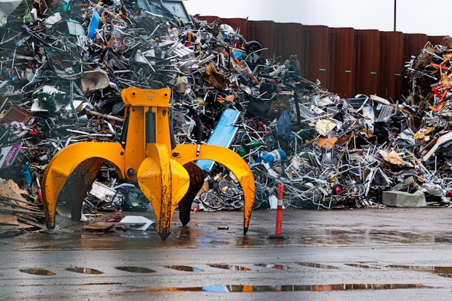 A large yellow mechanical claw is positioned in front of a massive pile of assorted metal scrap and debris. The ground is wet, suggesting recent rain, and the background shows a corrugated metal fence. Various pieces of metal, including bicycle parts and wires, are scattered and piled haphazardly.