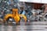 A large yellow mechanical claw is positioned in front of a massive pile of assorted metal scrap and debris. The ground is wet, suggesting recent rain, and the background shows a corrugated metal fence. Various pieces of metal, including bicycle parts and wires, are scattered and piled haphazardly.