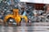 A large yellow mechanical claw is positioned in front of a massive pile of assorted metal scrap and debris. The ground is wet, suggesting recent rain, and the background shows a corrugated metal fence. Various pieces of metal, including bicycle parts and wires, are scattered and piled haphazardly.