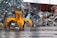 A large yellow mechanical claw is positioned in front of a massive pile of assorted metal scrap and debris. The ground is wet, suggesting recent rain, and the background shows a corrugated metal fence. Various pieces of metal, including bicycle parts and wires, are scattered and piled haphazardly.