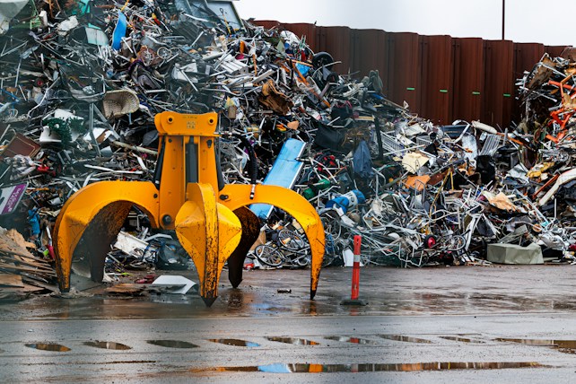 A large yellow mechanical claw is positioned in front of a massive pile of assorted metal scrap and debris. The ground is wet, suggesting recent rain, and the background shows a corrugated metal fence. Various pieces of metal, including bicycle parts and wires, are scattered and piled haphazardly.