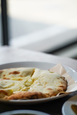 A close-up of a white plate containing a whole naan bread with a perfectly golden-brown crust, placed on a wooden table with a blurred background.