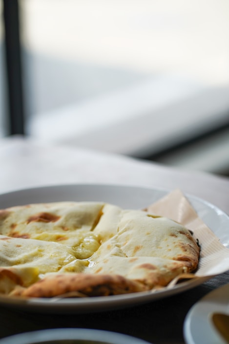A close-up of a white plate containing a whole naan bread with a perfectly golden-brown crust, placed on a wooden table with a blurred background.
