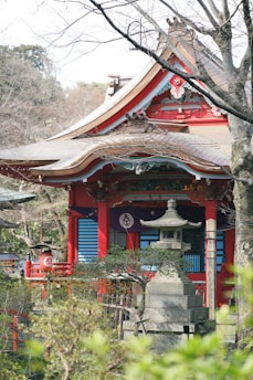 A traditional Japanese shrine features a vibrant red exterior with intricate architectural details. In front of the shrine, there is a stone lantern partially obscured by lush green foliage. Leafless trees surround the shrine, hinting at a serene and peaceful atmosphere.