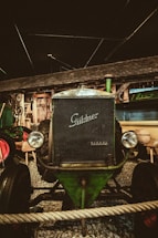 An antique Guldner Diesel tractor is displayed indoors against a rustic wooden backdrop. The tractor's vintage design features large round headlights and a prominent grill. It is surrounded by gravel and a rope barrier, enhancing its classic and preserved appearance.