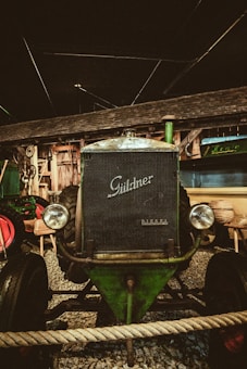 An antique Guldner Diesel tractor is displayed indoors against a rustic wooden backdrop. The tractor's vintage design features large round headlights and a prominent grill. It is surrounded by gravel and a rope barrier, enhancing its classic and preserved appearance.