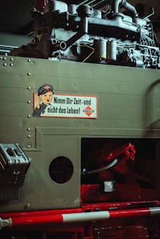 A vintage mechanical device is partially visible, featuring an old engine structure. A sign with a cartoon character and German text is attached to the machine, suggesting a safety message. The industrial components are predominantly metallic with visible rivets and mechanical parts.