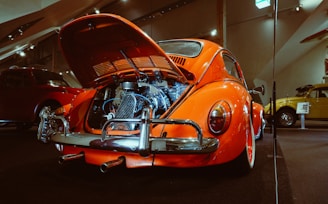 A vintage orange Volkswagen Beetle with its engine hood open, revealing the engine. The car is parked indoors in what seems to be an exhibit with other vintage cars also visible in the background. The setting is well-lit with overhead lights, and reflections can be seen on the polished surfaces of the cars.