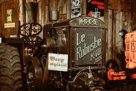 A vintage tractor with the name 'Le Robuste K40' on the front is positioned in what appears to be a rustic, possibly museum-like setting. The tractor is black with large, textured tires and has an old-fashioned steering mechanism. Several vintage posters and signs are visible in the background, enhancing the antique ambiance.
