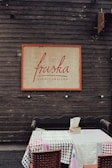 A wooden wall backdrop with a large framed sign reading 'fruska PICNIC & BISTRO' in red script. Below, a table is covered with a checked tablecloth in red, blue, pink, and white, with a wooden bench and chair adjacent to it.