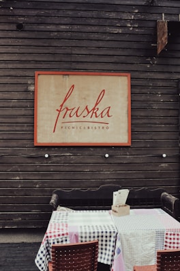 A wooden wall backdrop with a large framed sign reading 'fruska PICNIC & BISTRO' in red script. Below, a table is covered with a checked tablecloth in red, blue, pink, and white, with a wooden bench and chair adjacent to it.