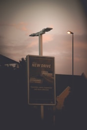 A tall street sign is illuminated against a moody evening sky, with two street lamps glowing softly nearby. In the background, silhouettes of trees and building structures are barely visible. The sign features advertising text focused on a new driving experience.