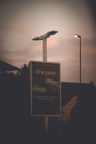 A tall street sign is illuminated against a moody evening sky, with two street lamps glowing softly nearby. In the background, silhouettes of trees and building structures are barely visible. The sign features advertising text focused on a new driving experience.