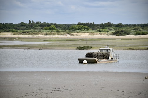 A flat boat loaded with oyster racks is parked in a shallow, muddy estuary. The landscape is serene, with sandy banks, some vegetation in the distance, and a cloudy sky.