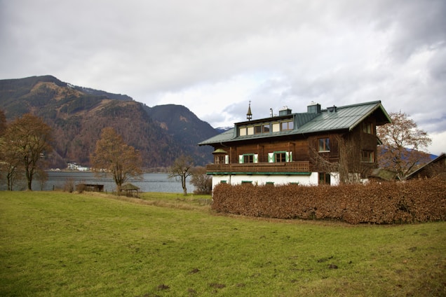 A large wooden house with a green roof is situated by a lake, surrounded by grassy fields and leafless trees. In the background, there are tall mountains under a cloudy sky.