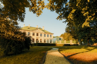 A historic mansion with classical architecture, featuring a green roof and large windows, is surrounded by lush, well-maintained gardens. A gravel pathway leads through the lawn toward the building, and the scene is bathed in warm, golden sunlight filtering through the trees.