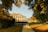 A historic mansion with classical architecture, featuring a green roof and large windows, is surrounded by lush, well-maintained gardens. A gravel pathway leads through the lawn toward the building, and the scene is bathed in warm, golden sunlight filtering through the trees.