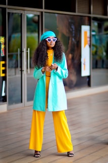 A person with long curly hair is dressed in a vibrant outfit, featuring a turquoise coat, matching beret, and yellow wide-leg pants. They wear stylish white sunglasses and black sandals, standing against a backdrop of glass doors and a reflective surface.