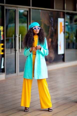 A person with long curly hair is dressed in a vibrant outfit, featuring a turquoise coat, matching beret, and yellow wide-leg pants. They wear stylish white sunglasses and black sandals, standing against a backdrop of glass doors and a reflective surface.