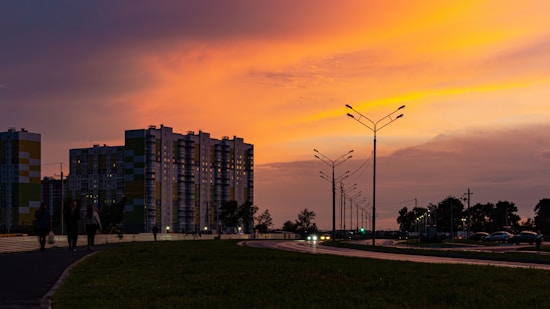High-rise apartment buildings stand against a vibrant sunset sky, with deep orange and pink hues. Streetlights line the road, illuminated in the early evening. There are silhouettes of people walking on the sidewalk, and trees are visible in the background.