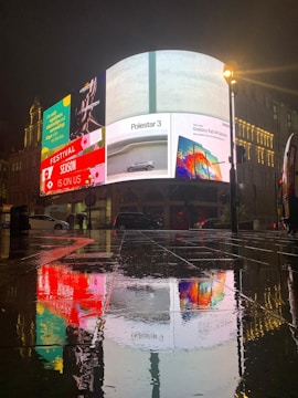 A large illuminated billboard dominates the scene, featuring advertisements for various brands. The wet pavement reflects the bright lights and colors, creating a vivid and dynamic visual effect. Surrounding the billboard are traditional-style buildings, and a few cars are visible on the street, indicating an urban setting.