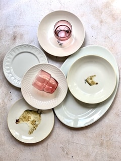 A collection of plates and bowls arranged on a textured surface. Two pink glasses sit on top of the dishes. Some of the plates feature a chicken illustration. The arrangement suggests a rustic or vintage style with an emphasis on simplicity.
