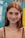A young woman with long red hair, freckles, and large hoop earrings smiles gently towards the camera. She wears a black spaghetti strap top and a delicate necklace, standing in front of a colorful background featuring various blurred logos and text.