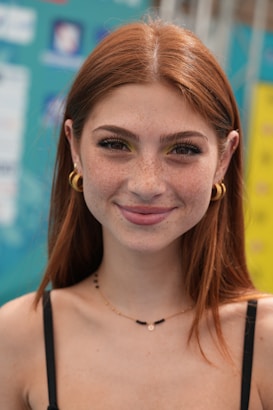 A young woman with long red hair, freckles, and large hoop earrings smiles gently towards the camera. She wears a black spaghetti strap top and a delicate necklace, standing in front of a colorful background featuring various blurred logos and text.
