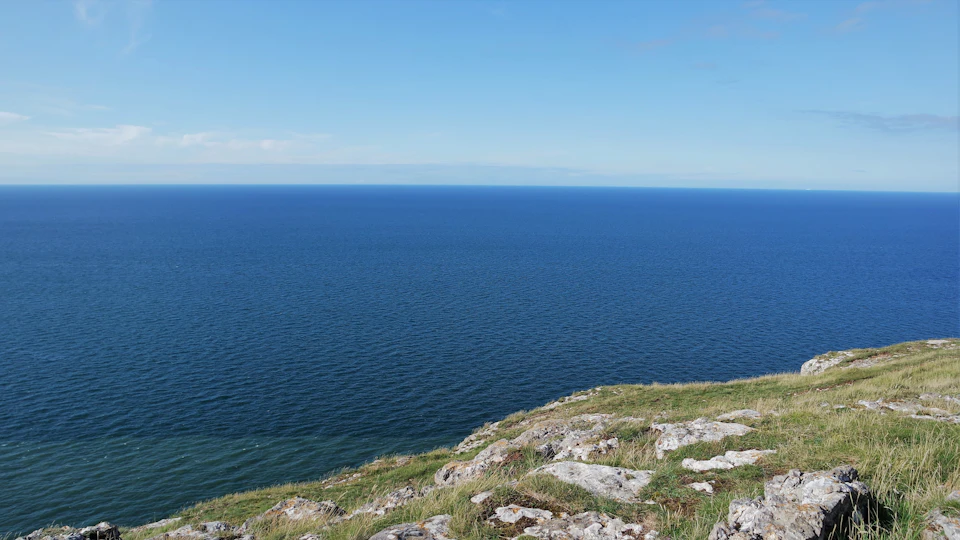 The image depicts a serene coastal landscape with a rocky cliff overlooking the ocean. The cliff is covered in green grass and features several large rocks scattered across its surface, adding texture to the scene. The vast expanse of the deep blue ocean stretches out towards the horizon, meeting the clear sky at the water's edge. The perspective from the top of the cliff provides an unobstructed view of this picturesque setting.